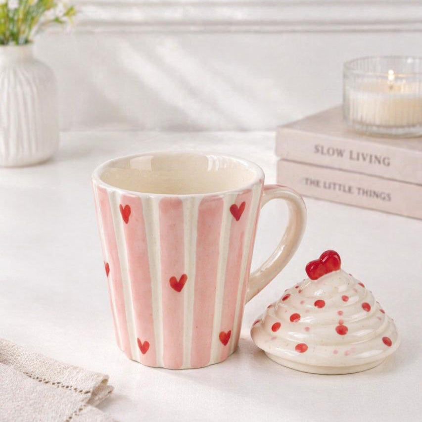 Pink mug with red heart patterns on a white surface with a small decorative item and books in the background.