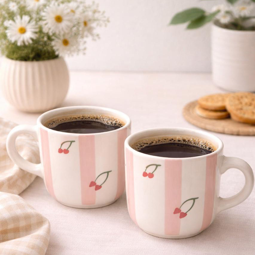 Two mugs with cherry design on a table with coffee and cookies.