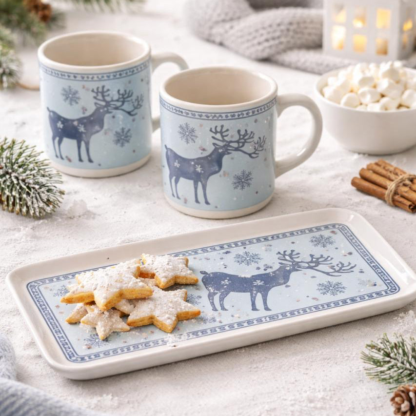 Two mugs with reindeer design on a tray with cookies, surrounded by festive decor.