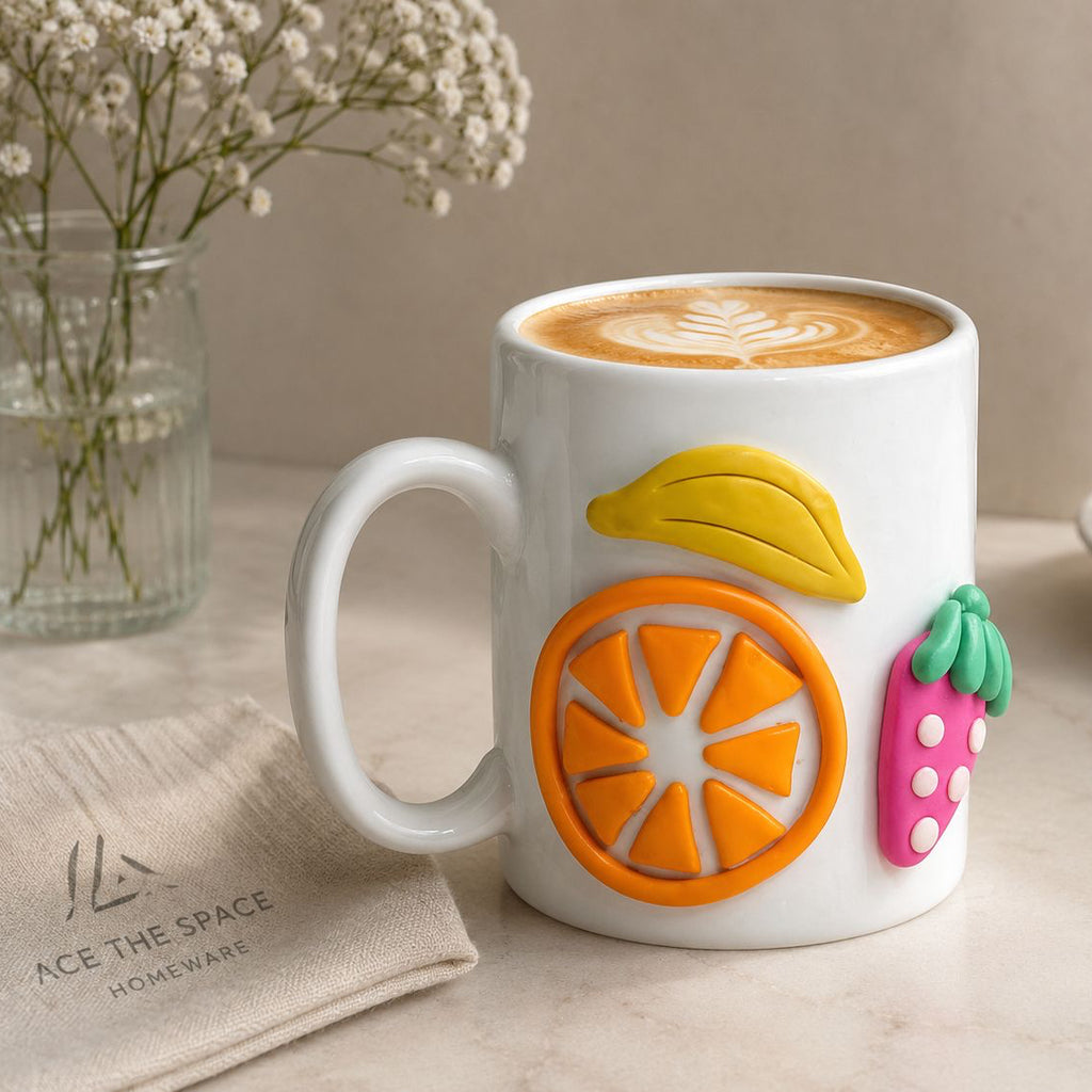 White mug with fruit designs filled with coffee, placed on a table with a plate of pastries and a vase of flowers.