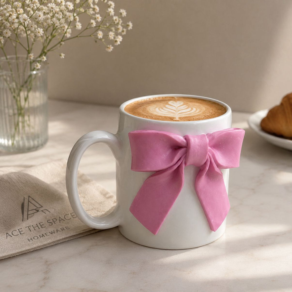 White mug with a pink bow, filled with coffee, on a marble surface with a croissant and flowers in the background.