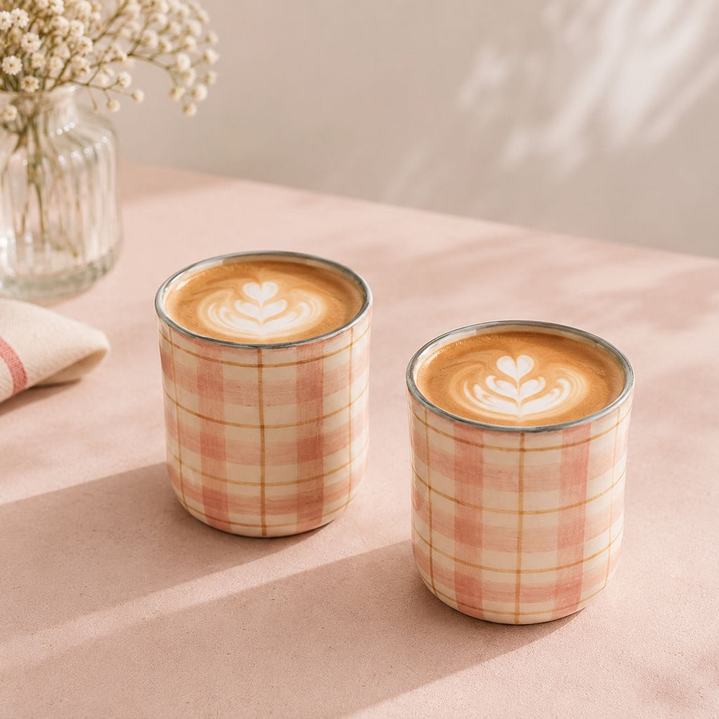 Two plaid-patterned coffee cups with latte art on a soft pink surface.