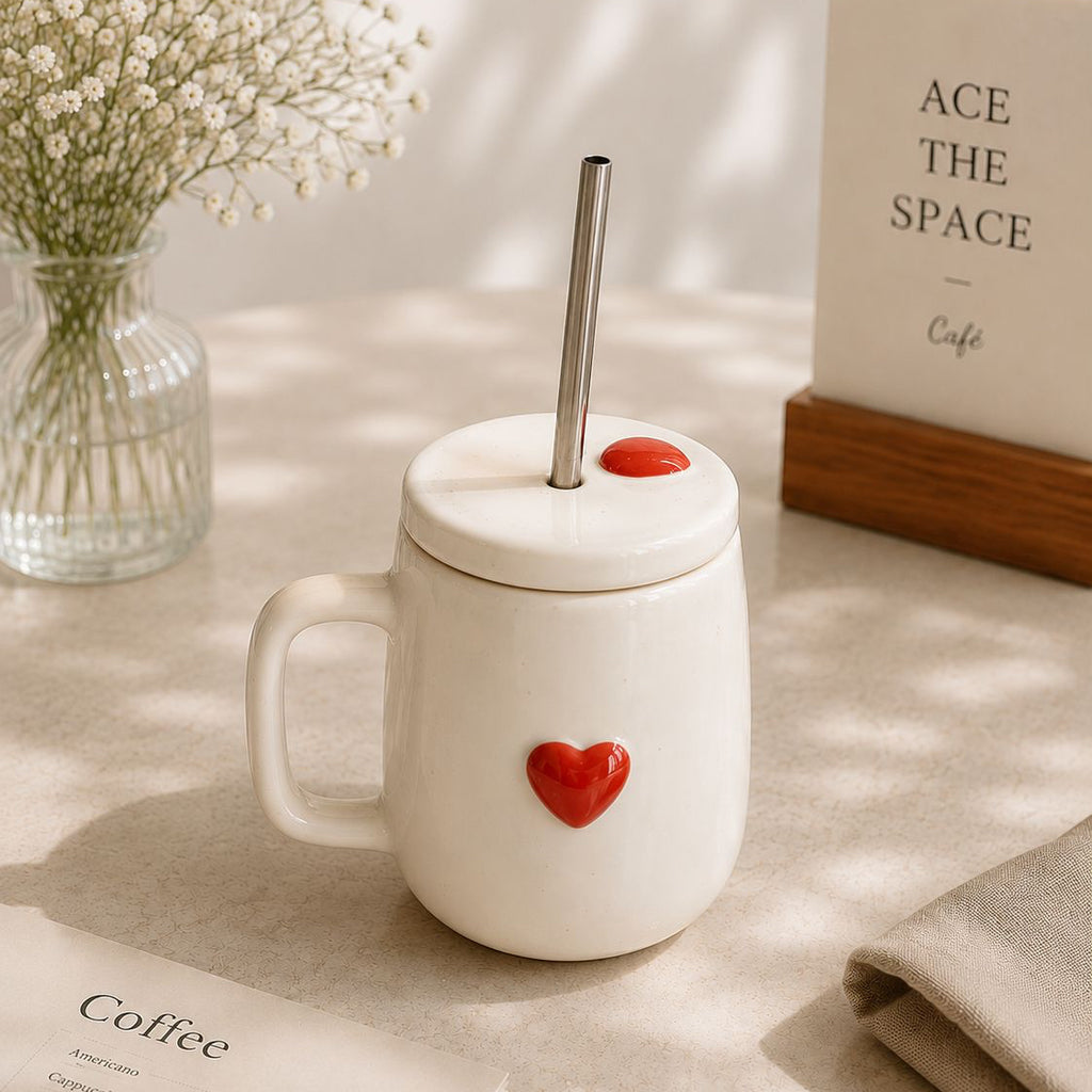 White mug with a red heart and straw lid on a table with a book and vase in the background