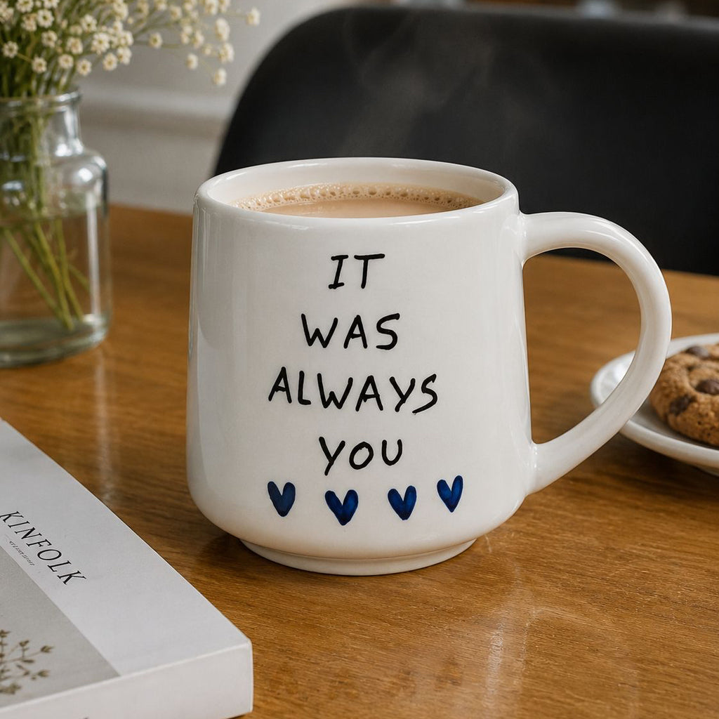 White mug with 'It was always you' text on a wooden table with a book and cookies.