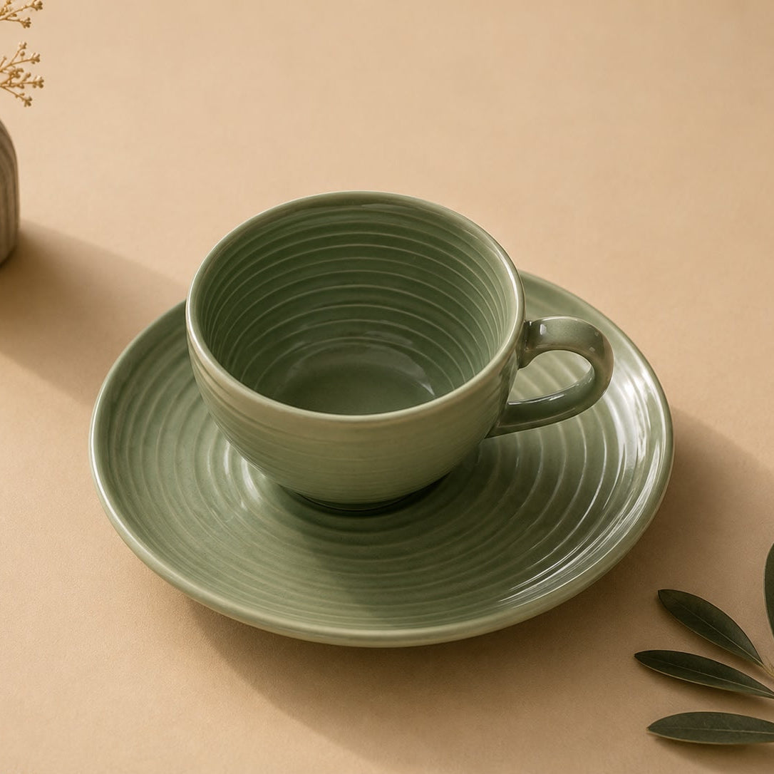 Green ceramic cup and saucer set on a beige surface with a small vase and leaves.
