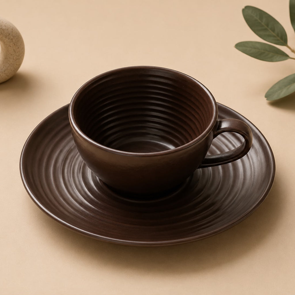 Brown ceramic cup and saucer set on a beige surface with a small vase and green leaves.