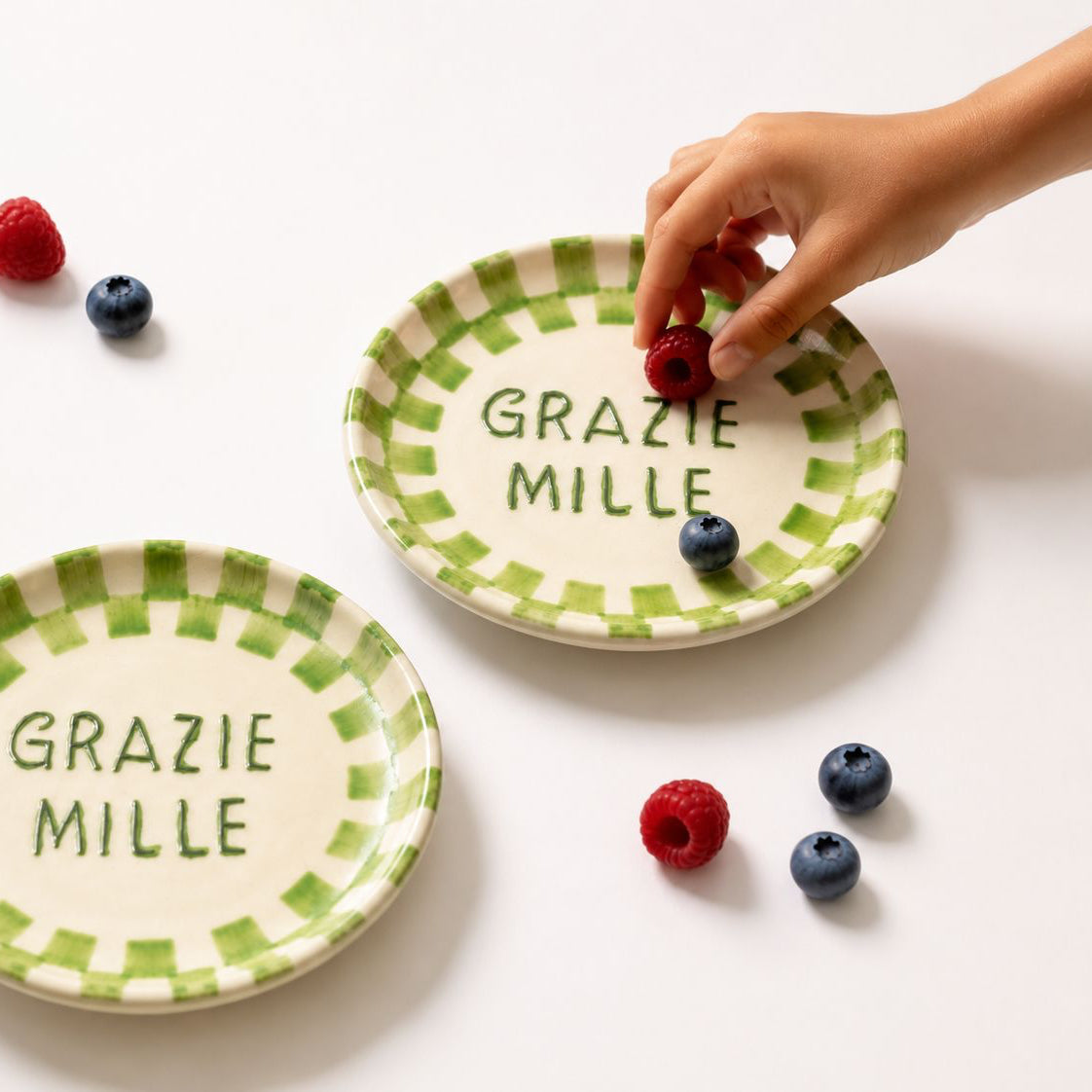 Two ceramic plates with 'Grazie Mille' text and checkered border, surrounded by berries on a white background.