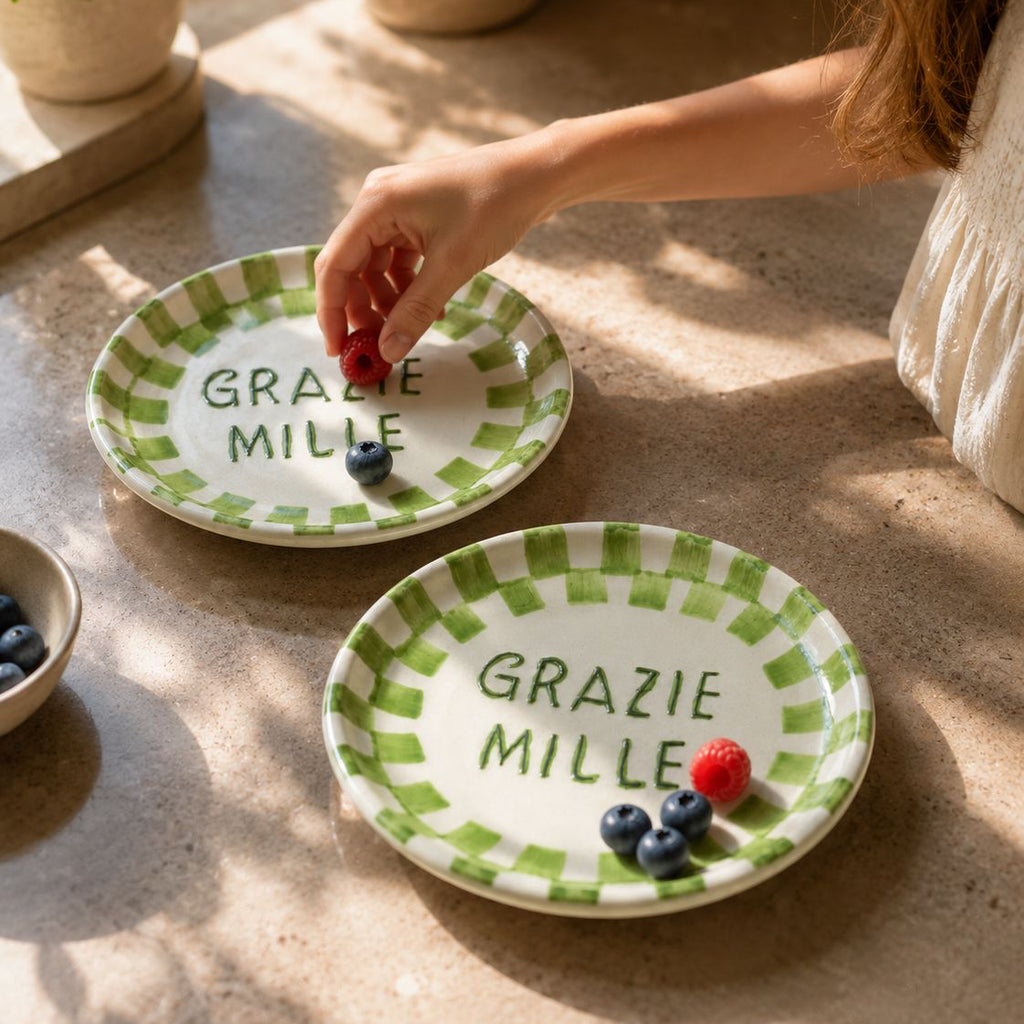 Two small ceramic plates with green checkered rim and 'Grazie Mille' text on a stone surface.