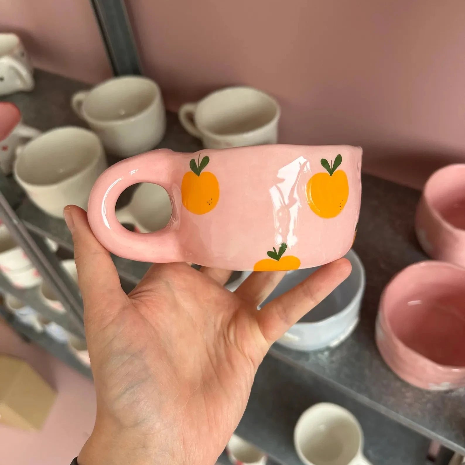 Hand holding a pink mug with orange designs against a background of more mugs.