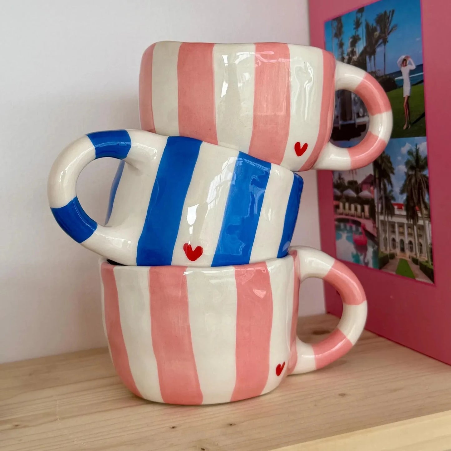 Stack of three ceramic mugs with pink and white stripes, blue stripes, and red hearts on a wooden surface.
