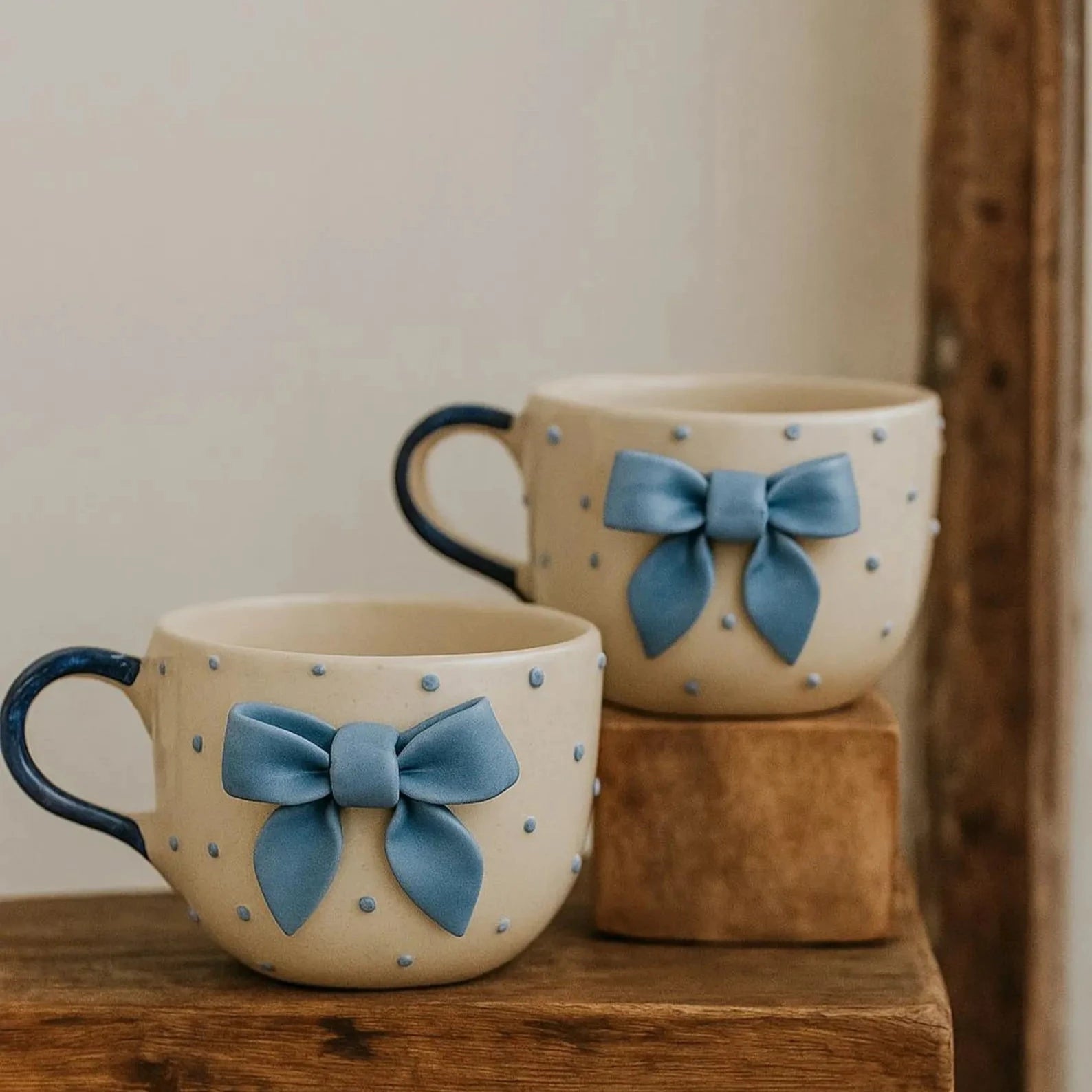 Two ceramic mugs with blue bow decorations on a wooden surface.